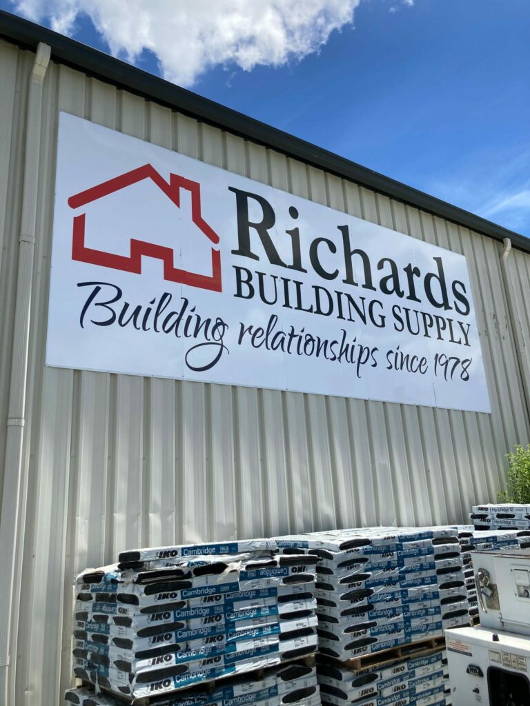 Richards Building Supply logo, black type and a red house on the outside of a beige metal building in front of a stack of shingles, Jacksonville, NC