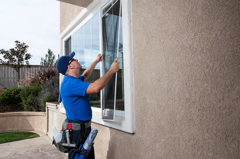 Man dressed in a blue t-shirt and blue hat, cleaning windows and screens on the exterior of a beige stucco home