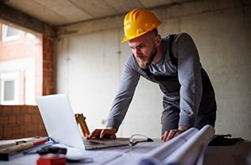 Construction worker with a hard hat looking at a laptop and blue prints